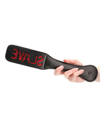 A hand gripping a plush black paddle with the word 'SLAVE' embossed in red, emphasising its tactile elegance and assertive design.