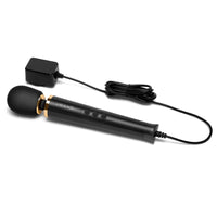 Le Wand Powerful Petite Plug-In Vibrating Massager displayed on a flat surface with the charging cord connected, emphasising its stylish black finish and gold accents.